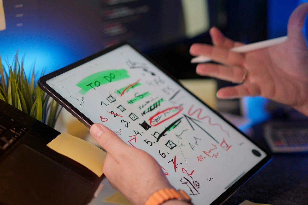 A person holding a tablet displaying a to-do list with various tasks and scribbles, some items are highlighted in green or circled in red.